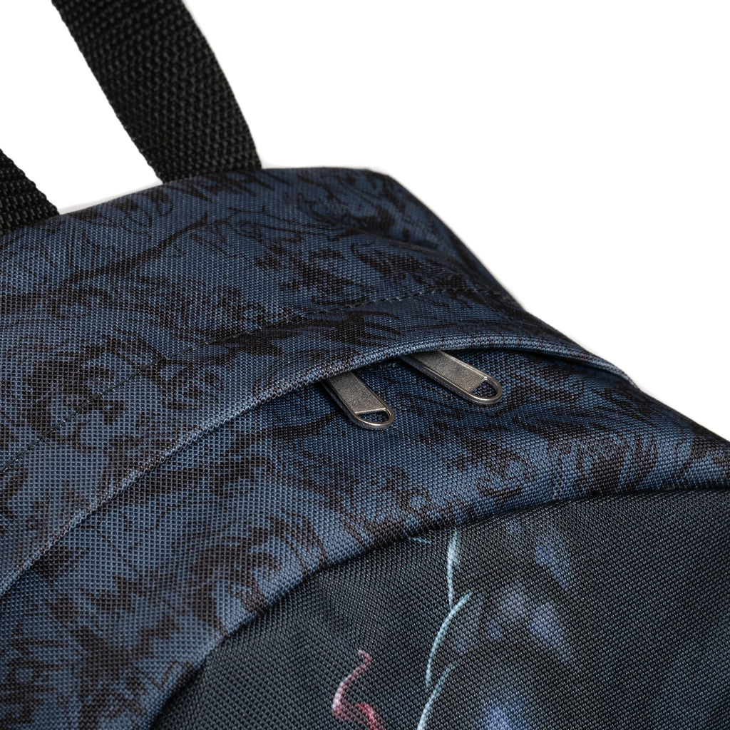 Close-up of the top of a dark charcoal backpack with a subtle black pattern, featuring a zippered pocket with silver metal zippers and a black webbing straps top handle, set against a plain white background.