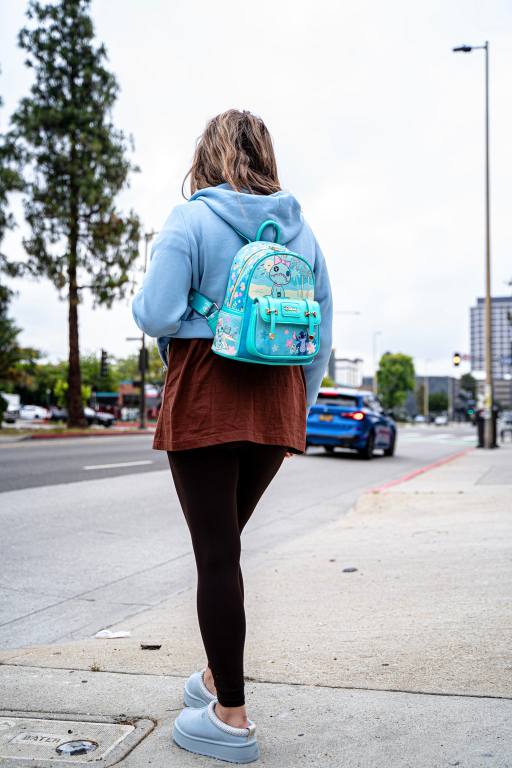 Woman walking on sidewalk near West Broadway building wearing a blue Disney Stitch-themed mini backpack featuring floral accents and character Voodoo doll.