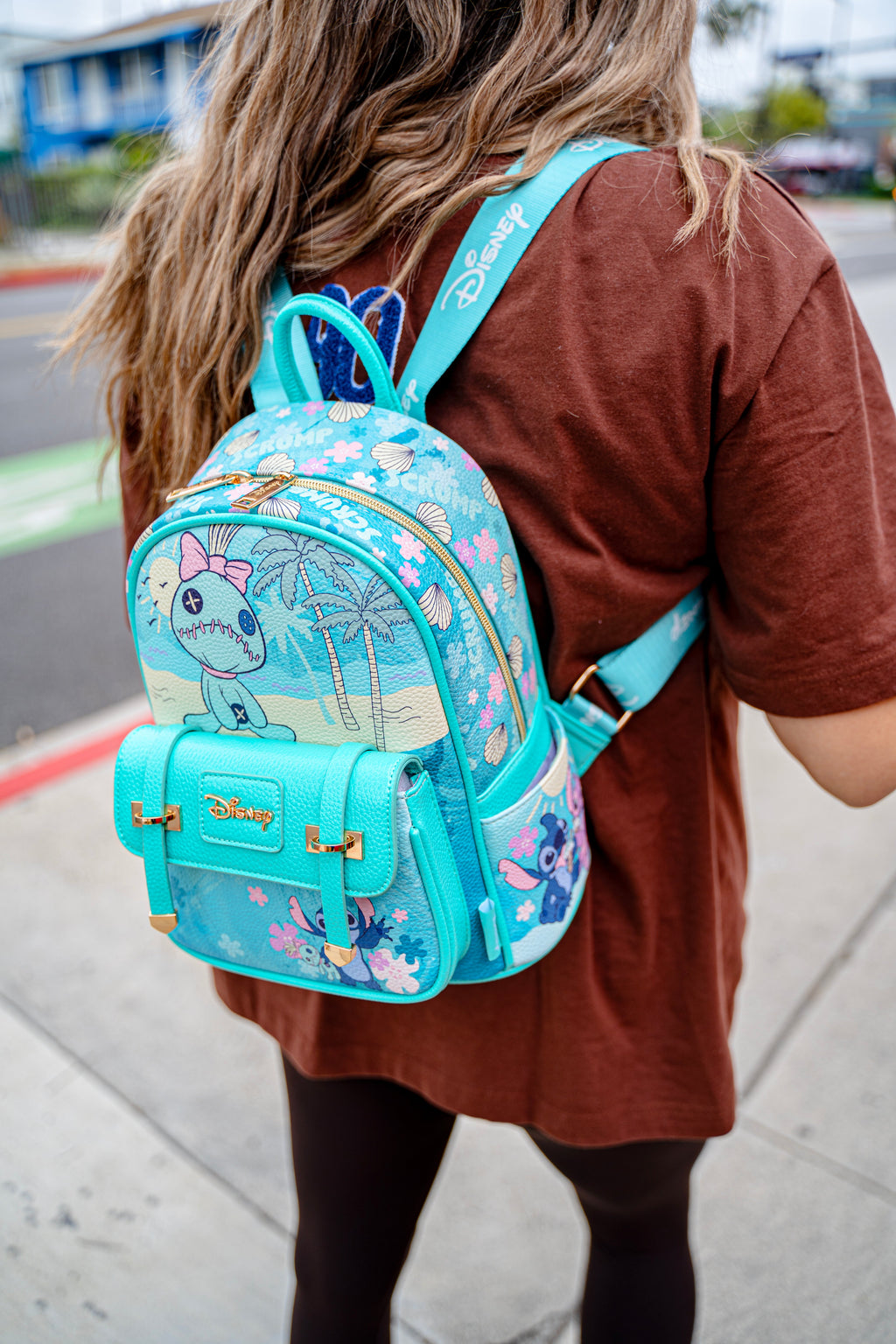 Zoomed-in view of a woman in a brown shirt wearing a bright blue Disney Stitch backpack with embroidered palm trees, flowers, and Disney branding on strap.