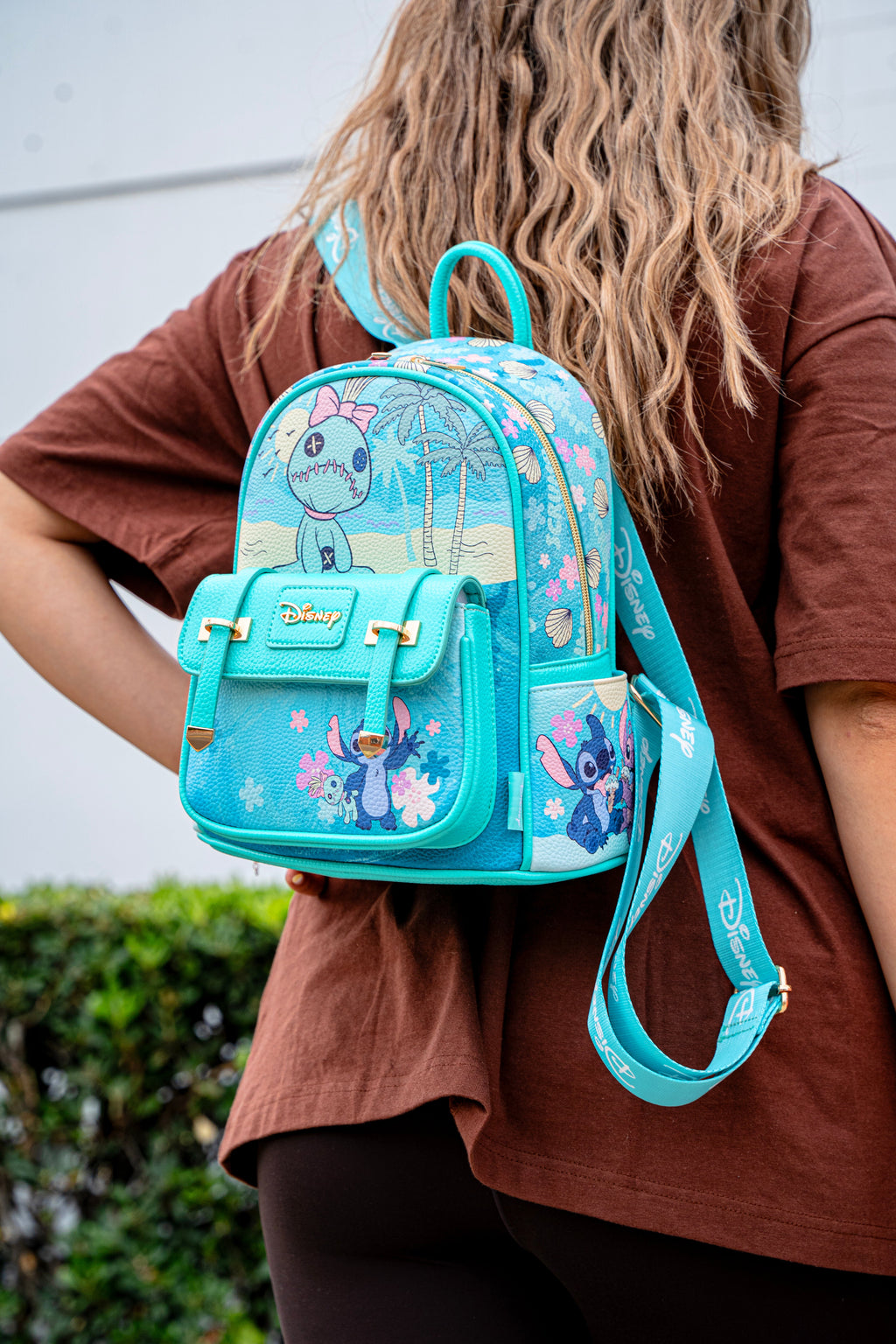 Close-up rear view of a woman in a blue hoodie and brown tunic showcasing a vibrant turquoise Disney Stitch mini backpack with palm trees and floral patterns.