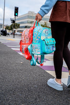 Alt Text:
Woman holding Stitch and Bambi mini backpacks by their handles while walking across a crosswalk, displaying detailed Disney character prints and colorful branded straps.